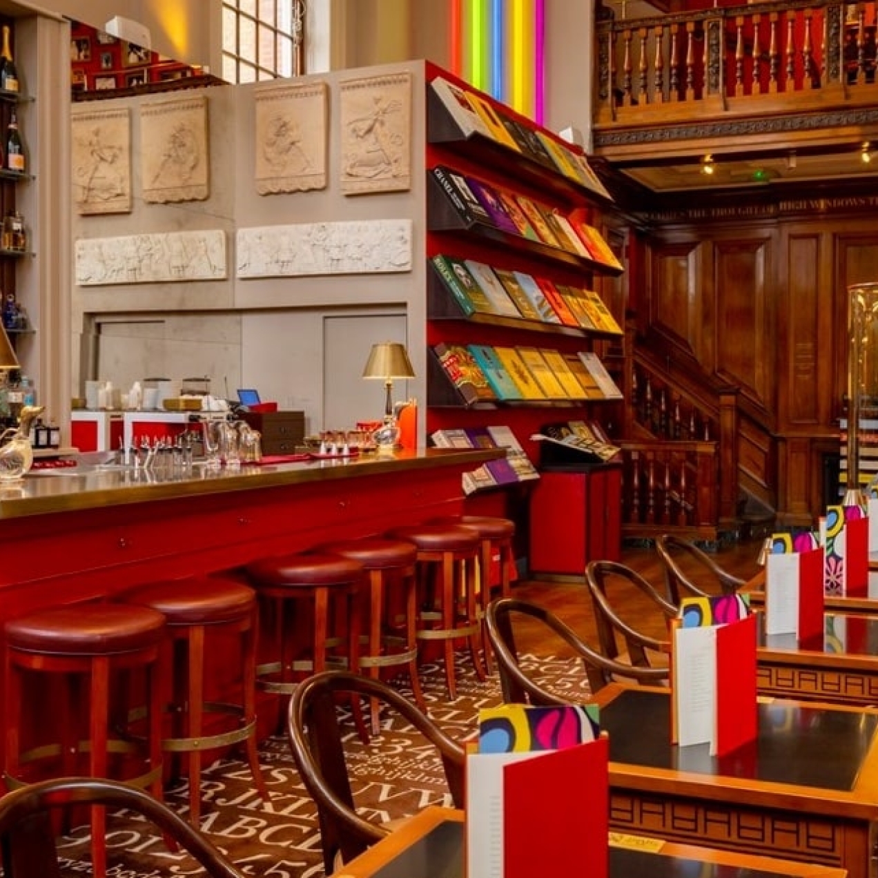 The interior of the Maison Assouline, with tables and chairs, menus, a bar with stools around it and lamps on top, and shelves full of bottles and books in the background